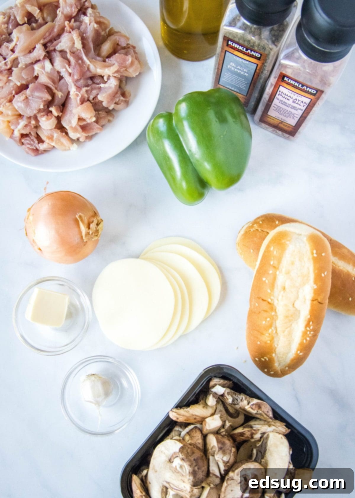 Overhead view of the ingredients you need for chicken Philly cheesesteaks: a bowl of sliced chicken, a green bell pepper, two hoagie rolls, a tub of mushrooms, an onion, slices of provolone cheese, a bowl of garlic, a bowl of butter, a jar of olive oil, a container of salt, and a container of pepper