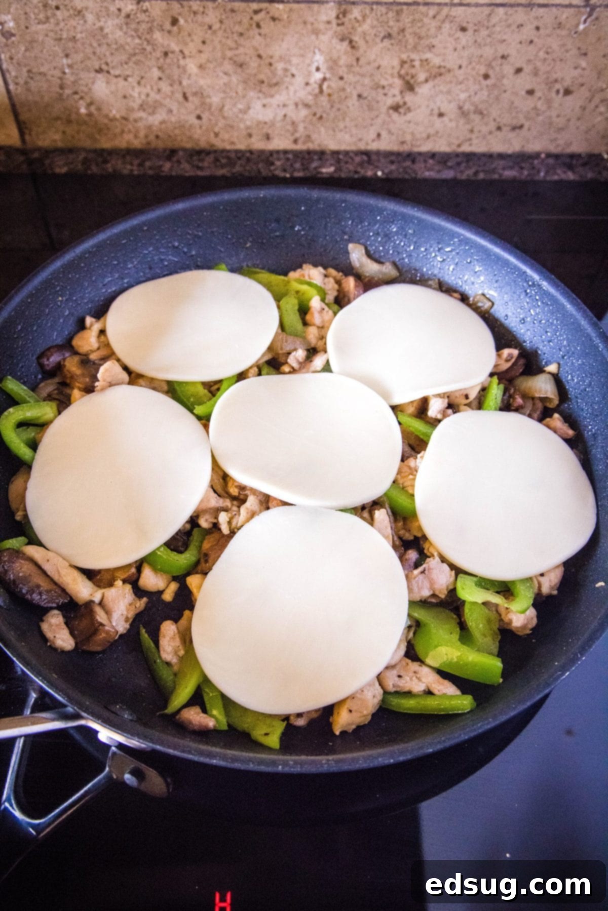 Slices of provolone cheese on top of onions, peppers, mushrooms, and chicken in a skillet