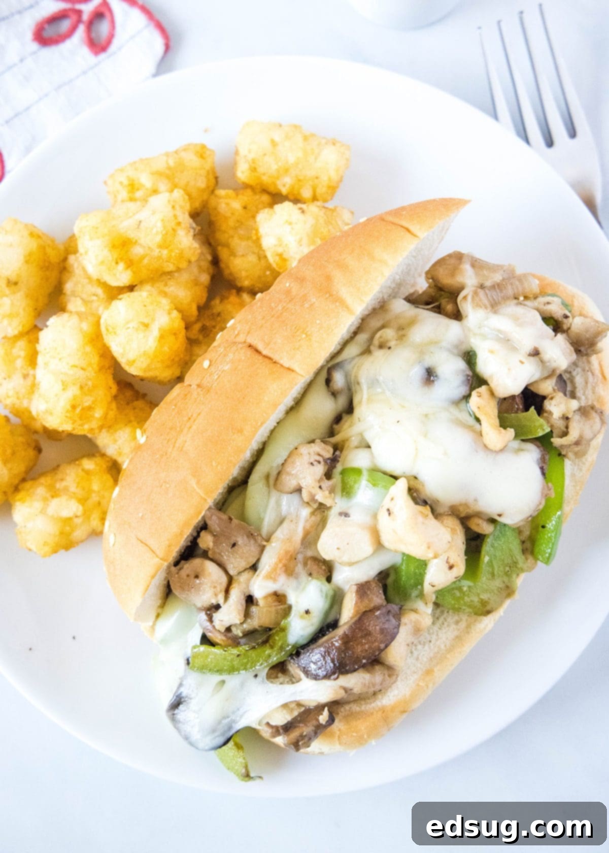 Overhead view of a chicken Philly cheesesteak on a plate next to some tater tots, with a fork on the side