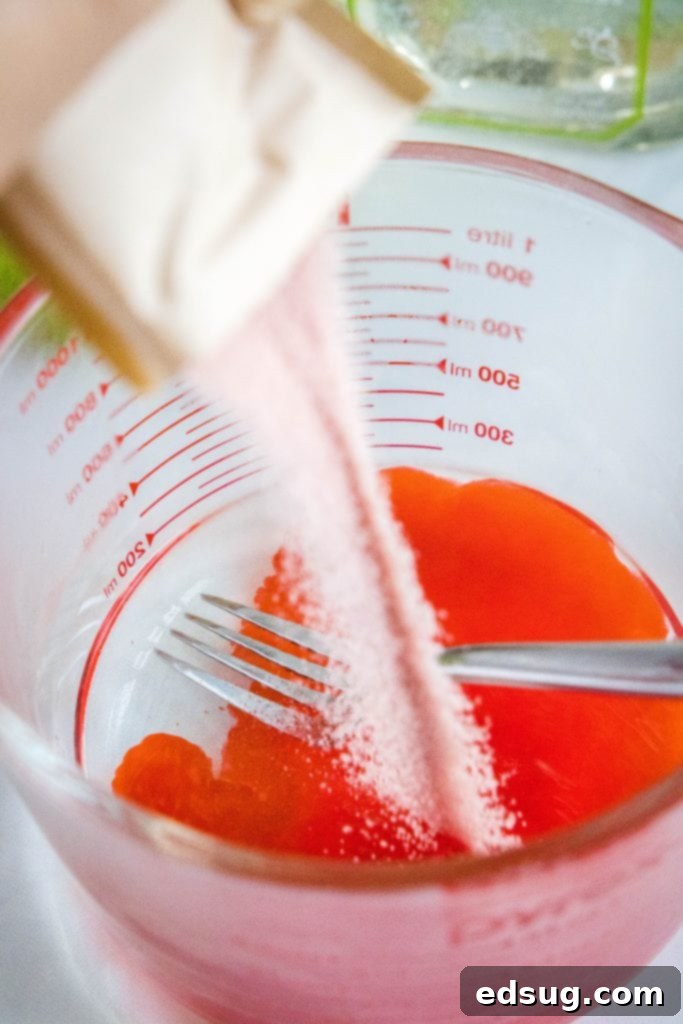 A hand pouring strawberry jello mix into a bowl of hot water, illustrating the first step of making jello shots.