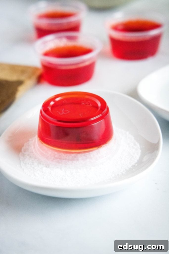 A hand coating the rim of a set strawberry margarita jello shot cup with coarse salt.