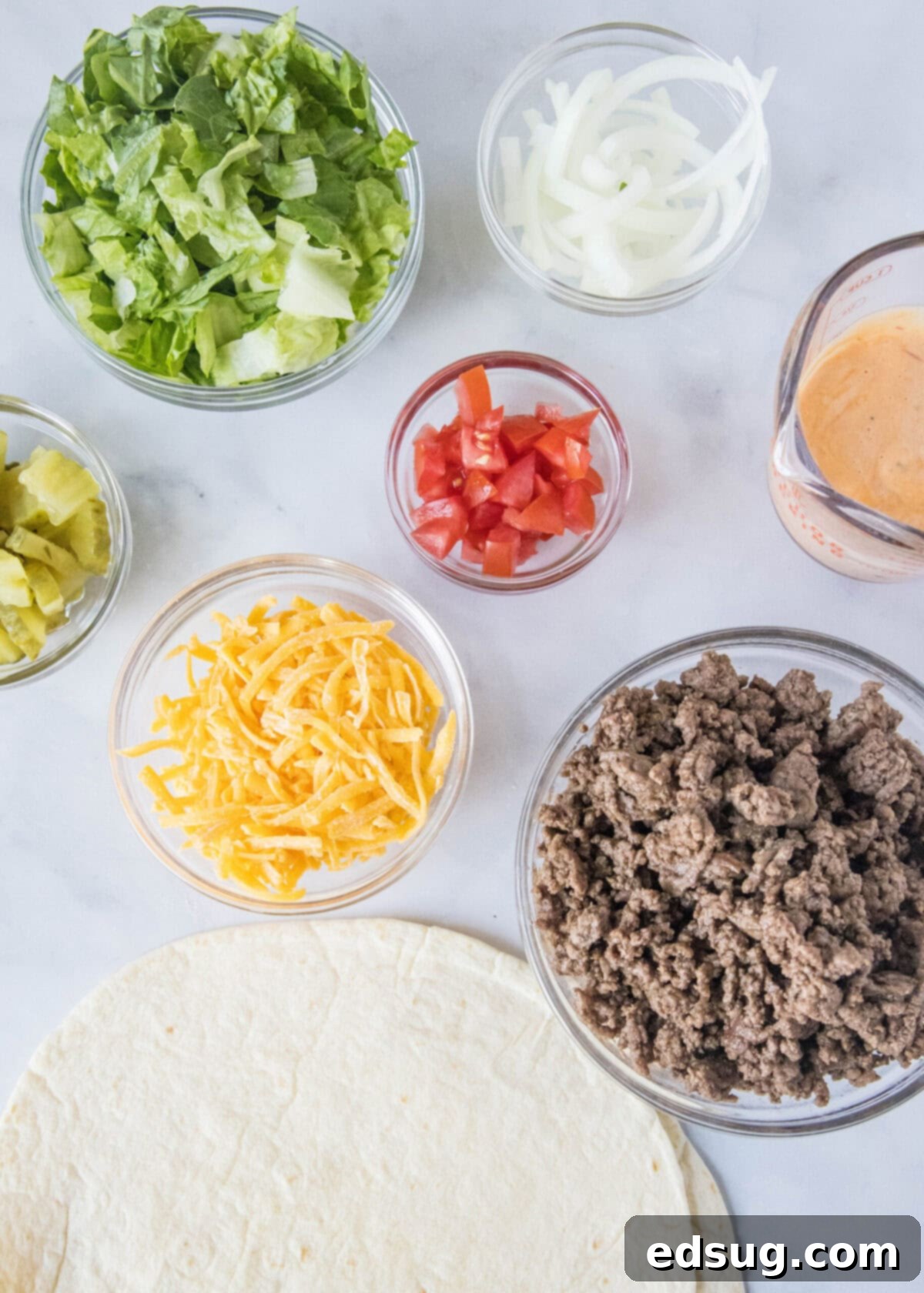 Overhead view of the ingredients needed for Big Mac wraps: a stack of tortillas, a bowl of cooked ground beef, a bowl of shredded cheese, a bowl of onions, a bowl of tomatoes, a bowl of pickles, a bowl of lettuce, and a jar of sauce