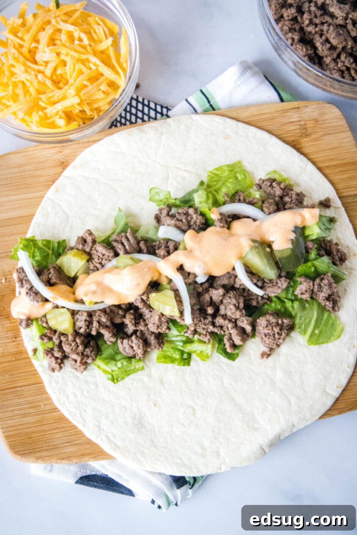 Overhead view of a flat tortilla topped with veggies, ground beef, and sauce, next to a bowl of cheese