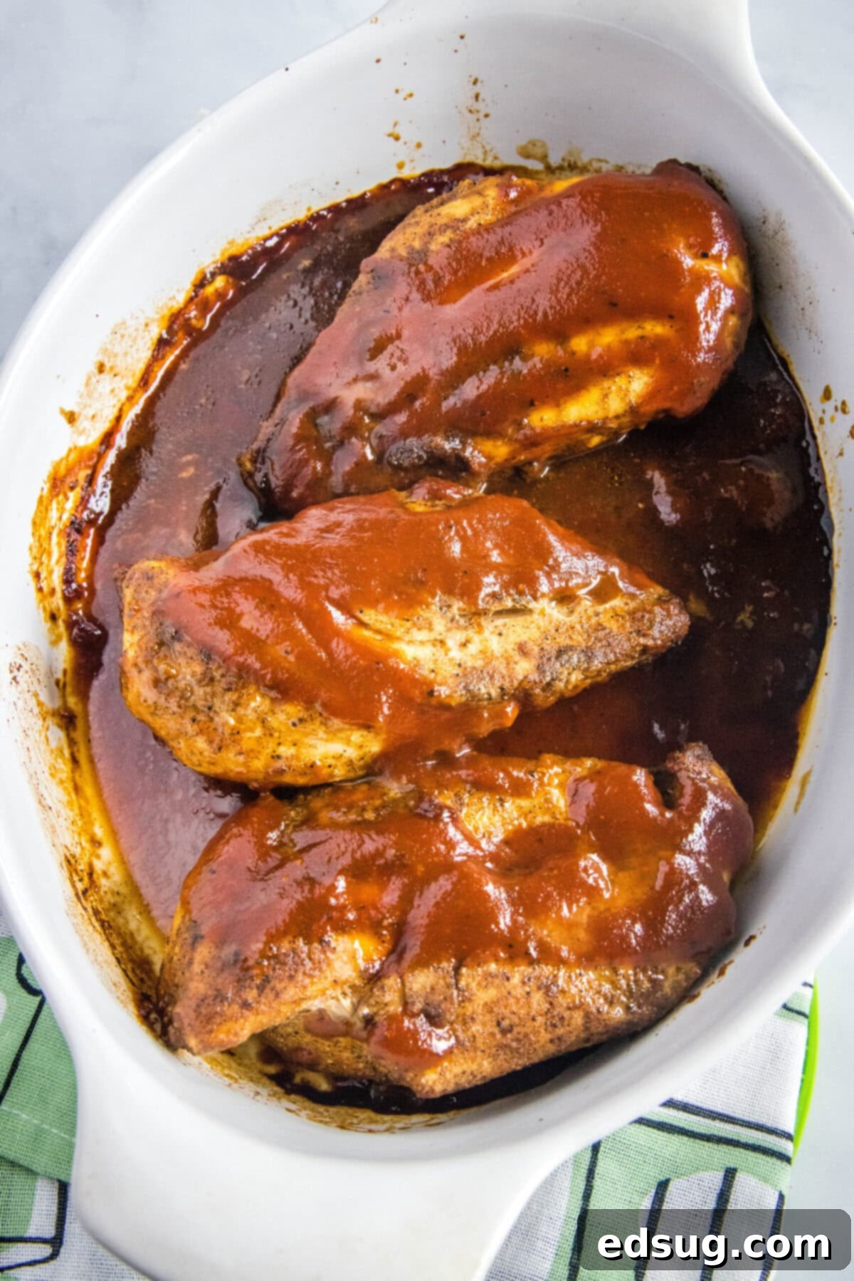 Overhead view of three chicken breasts fully covered in BBQ sauce in a baking dish, glistening and ready for final baking.