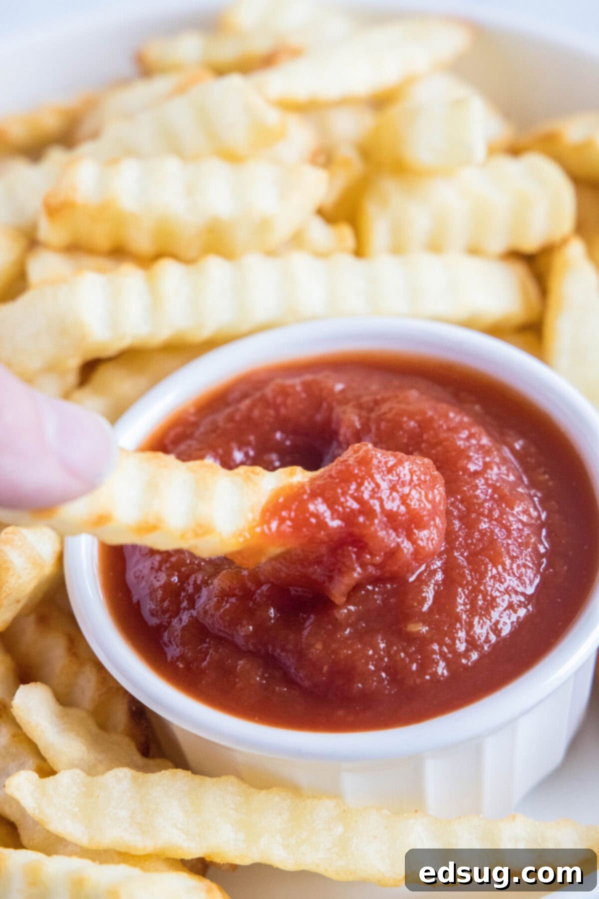 Homemade ketchup is tangy, salty, smooth, and so much better than the store-bought kind. Plus, it's easy to make and takes less than an hour. A hand dipping a crispy golden French fry into a ramekin of homemade ketchup, surrounded by a generous pile of fries.
