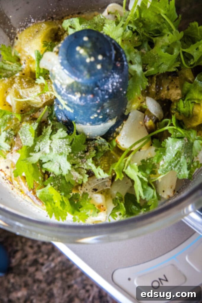 Roasted tomatillos and other vegetables being poured from a baking sheet into a food processor, with fresh cilantro added on top, ready for blending into smooth salsa verde.