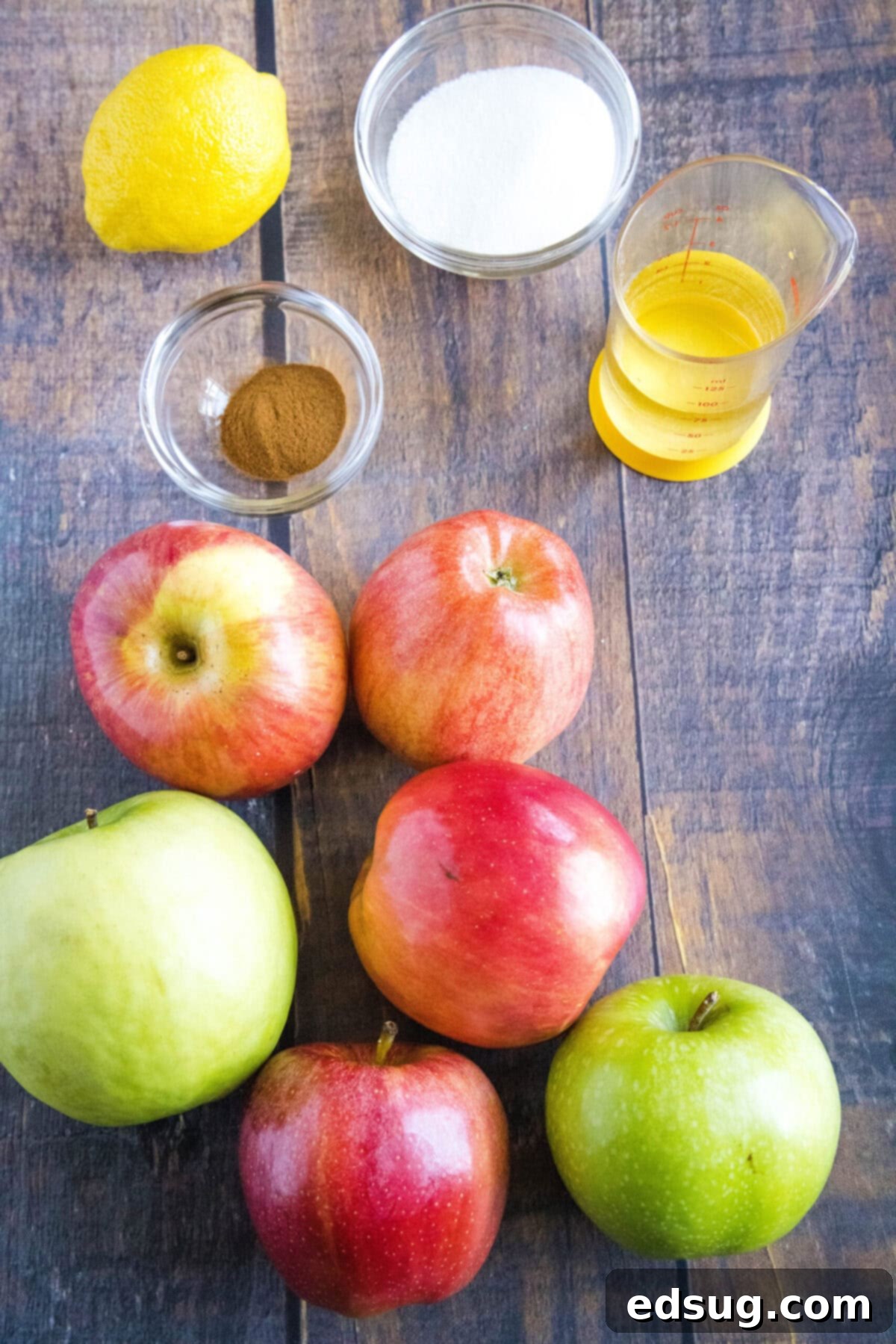 This homemade applesauce recipe is tart, lemony, not too sweet, bursting with apple flavor, and only takes 40 minutes to make.  Overhead view of the ingredients needed for homemade applesauce: a variety of apples, a lemon, a bowl of cinnamon, a bowl of sugar, and a glass of water