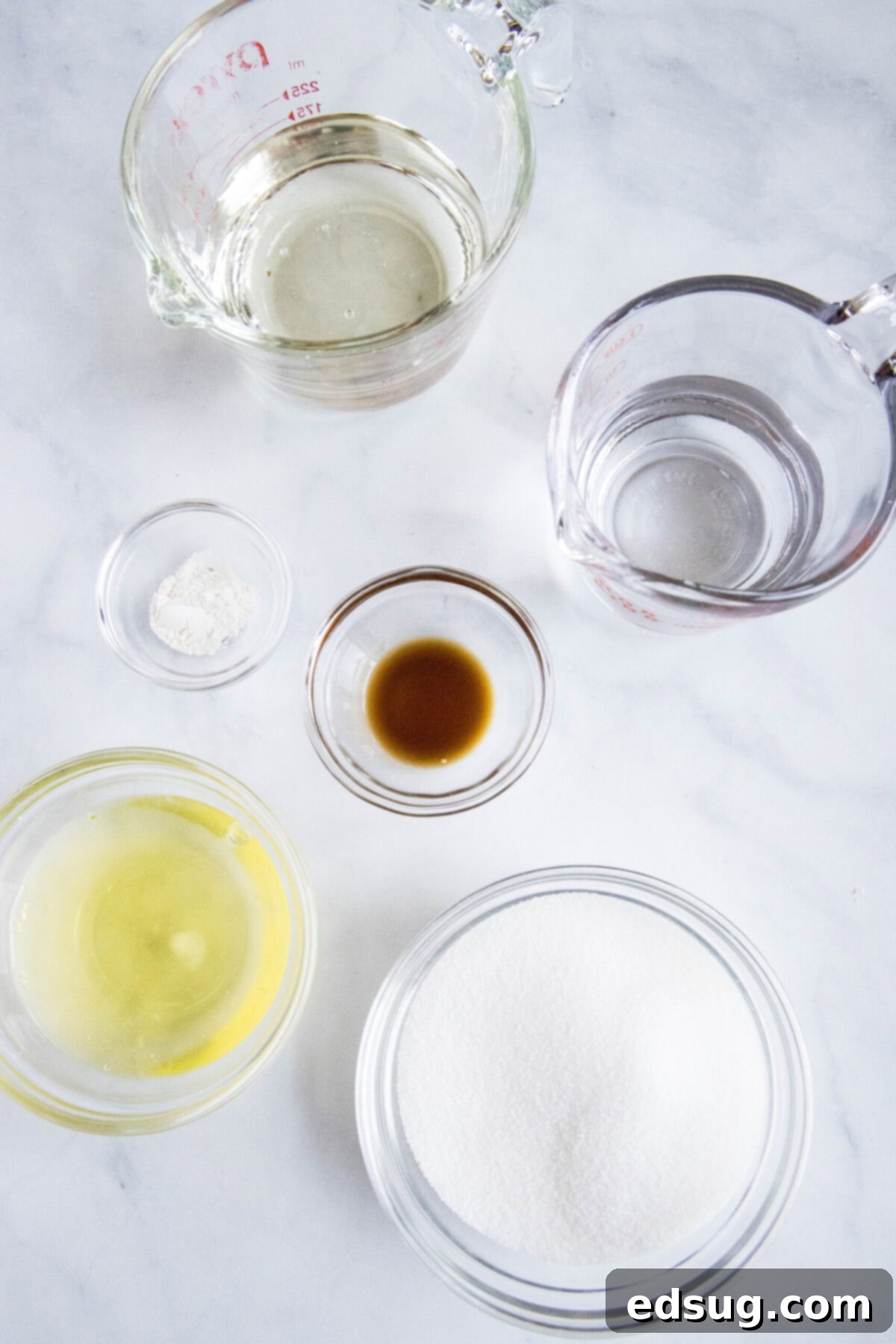 Overhead view of the ingredients needed for marshmallow fluff: a bowl of egg whites, a bowl of sugar, a pyrex of water, a bowl of vanilla, a pyrex of corn syrup, and a bowl of cream of tartar