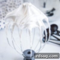 Close up of a stand mixer whisk attachment with stiff peaks of marshmallow fluff, with measuring spoons in the background