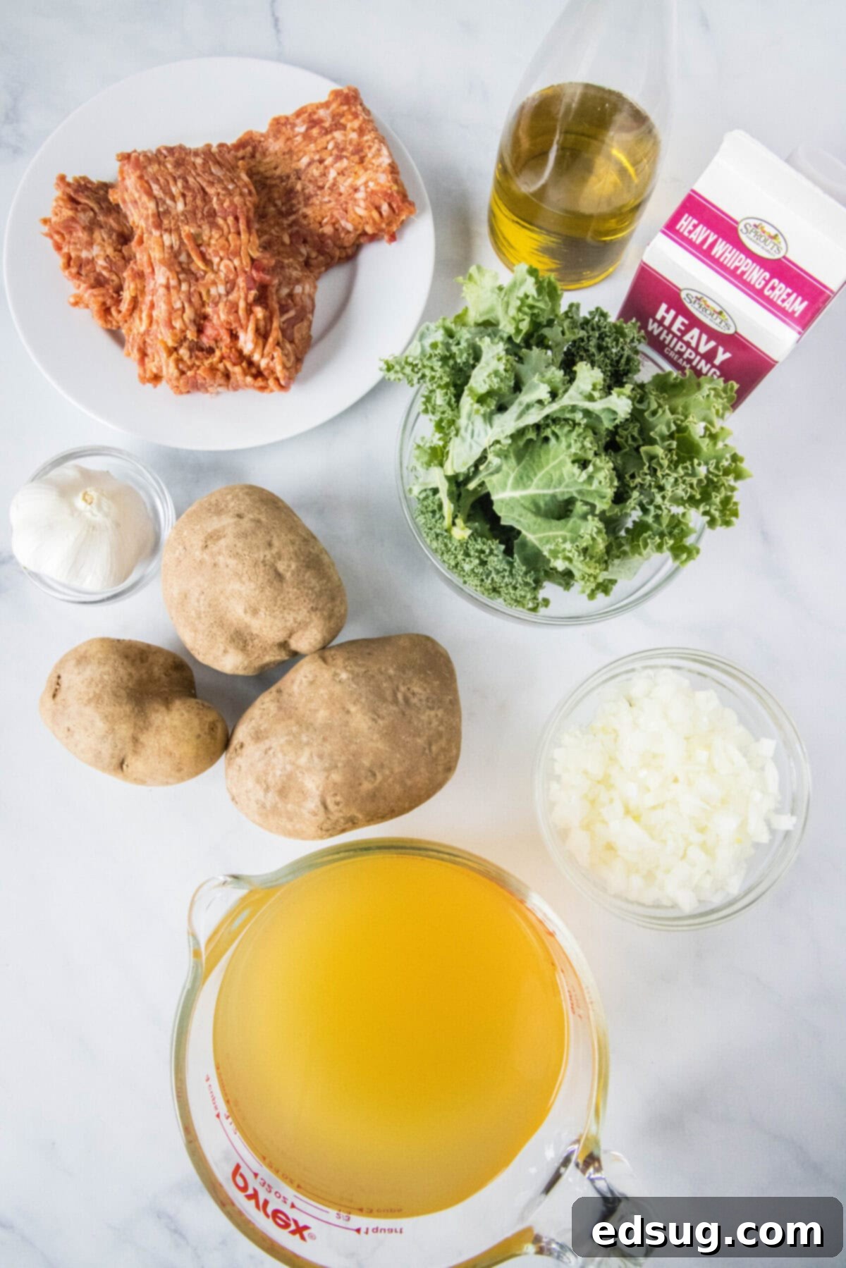 This copycat Olive Garden Zuppa Toscana is rich, hearty, healthy, loaded with potatoes and sausage, and easy to make in the Instant Pot. Overhead view of the ingredients needed for Zuppa Toscana: a plate of Italian sausage, a bowl of kale, a bowl of minced onion, a pyrex of chicken broth, a bowl of garlic, three potatoes, a jug of olive oil, and a carton of cream