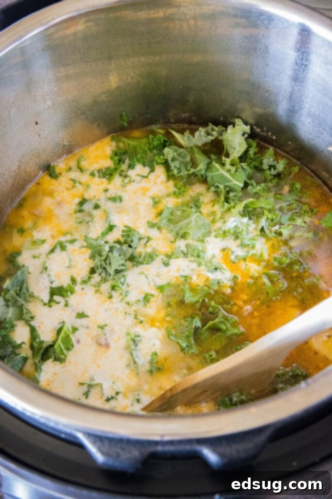 A wooden spoon stirring kale and cream into a pot of Zuppa Toscana