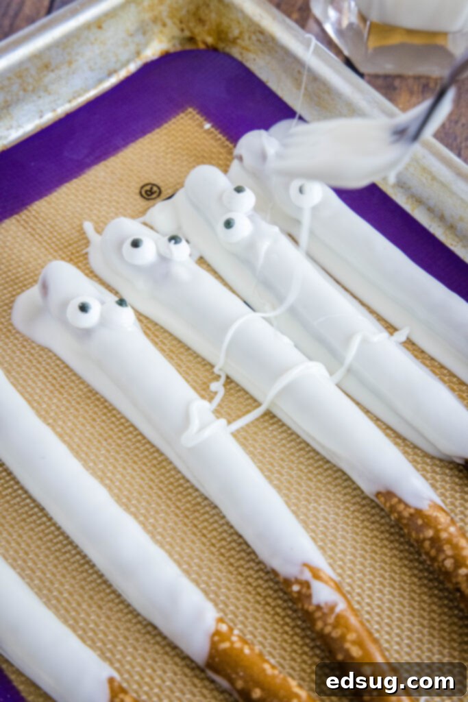 Bandaged Bites 5 A hand expertly drizzling melted white chocolate over decorated pretzel rods, forming the mummy bandage effect.
