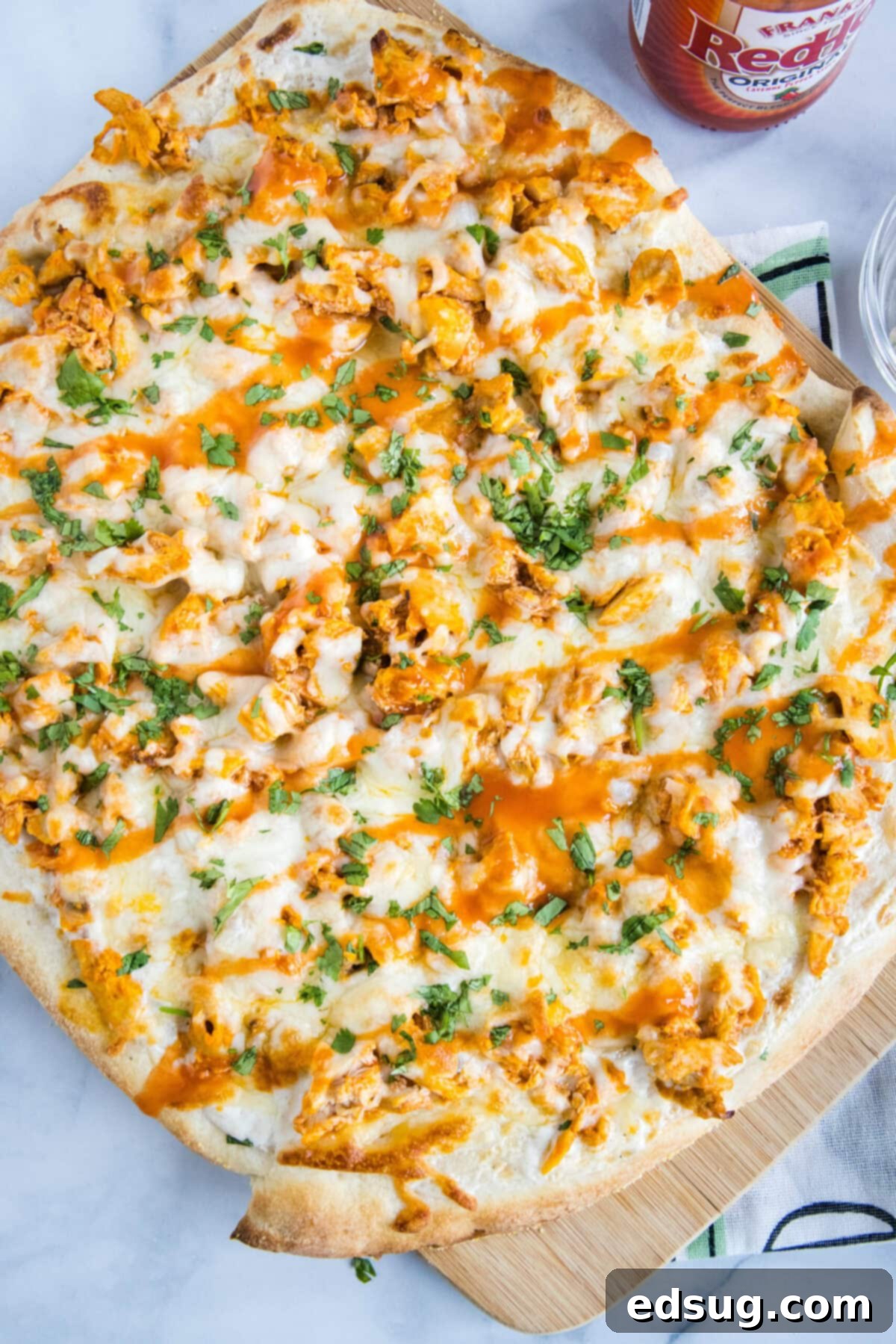 Overhead view of a whole buffalo chicken pizza next to a bottle of wing sauce, with a golden-brown crust, melted mozzarella, and spicy chicken visible