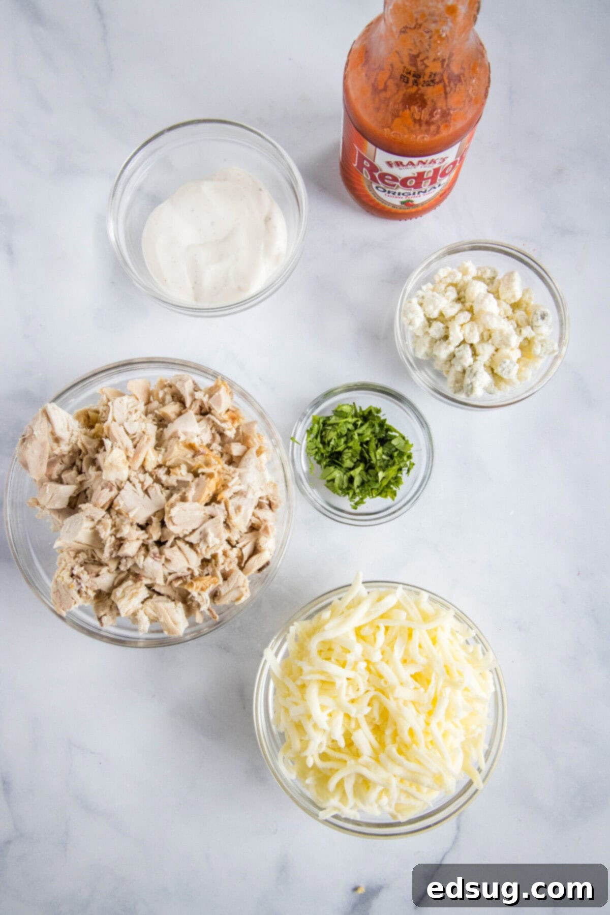 Overhead view of the fresh ingredients needed for buffalo chicken pizza: a bowl of shredded chicken, a bowl of shredded mozzarella cheese, a bowl of creamy ranch dressing, a bowl of blue cheese crumbles, a bowl of chopped parsley, and a bottle of bright red wing sauce.