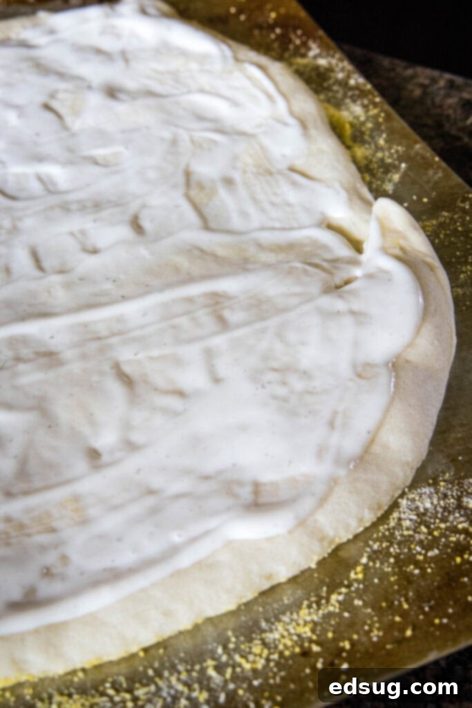 Creamy ranch dressing spread over a partially baked pizza dough, resting on a cornmeal-covered baking sheet, ready for toppings.