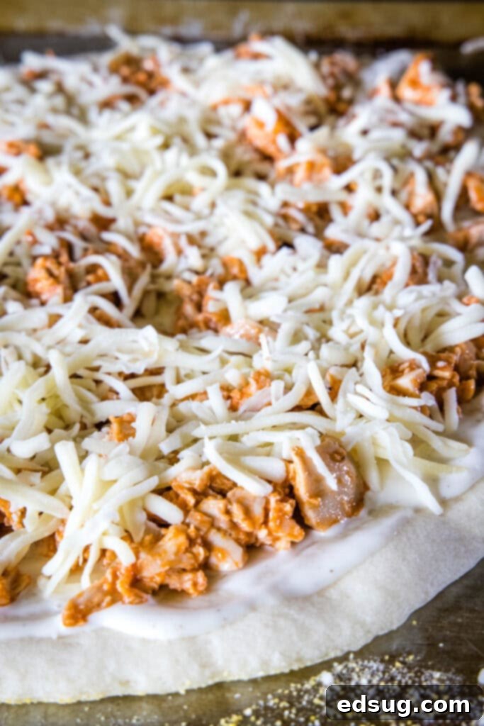 A buffalo chicken pizza assembled with spicy chicken and a layer of shredded mozzarella cheese on top of ranch dressing and a pre-baked crust, before final baking.