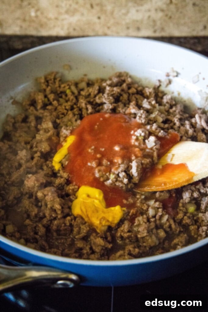The rich mixture of homemade sloppy joes in a skillet, showcasing tangy mustard and onions with savory ground beef. Adding yellow mustard and Worcestershire sauce on top of beautifully cooked ground beef and onions in a skillet, with a wooden spoon poised for stirring.