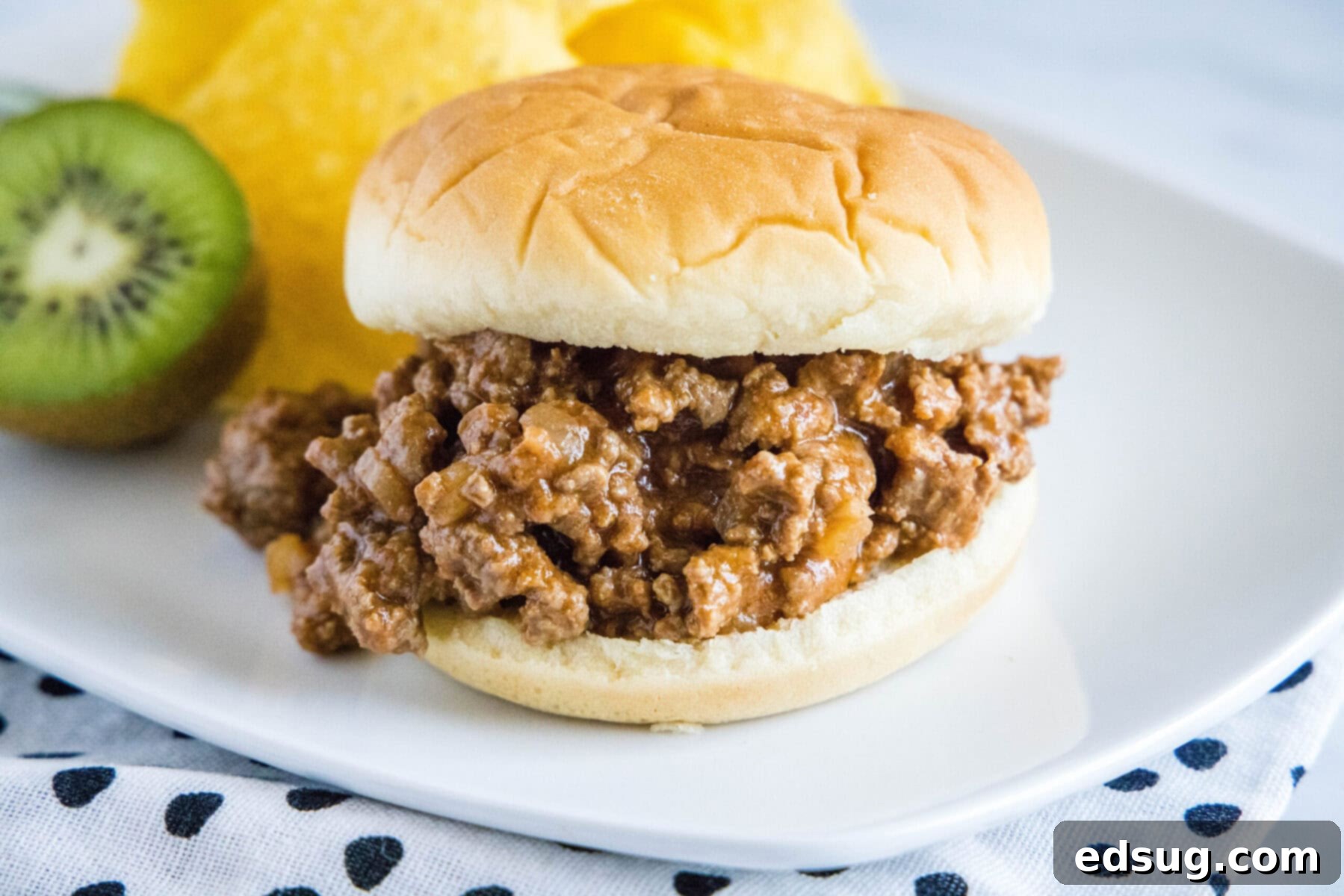 A delicious sloppy joe sandwich plated with a side of chips and a fresh kiwi, making for a wholesome and appealing meal. A perfectly assembled sloppy joe sandwich on a white plate, accompanied by a handful of crunchy potato chips and a vibrant slice of kiwi.