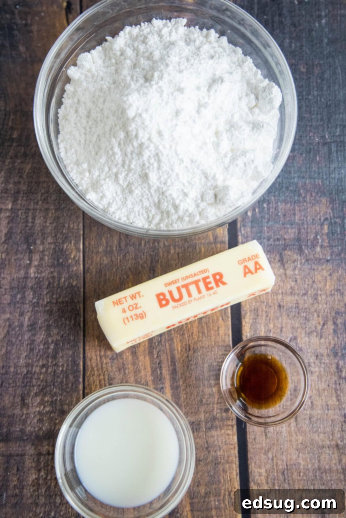 Overhead view of the ingredients needed for vanilla frosting: a bowl of powdered sugar, a bowl of milk, a bowl of vanilla, and a stick of butter