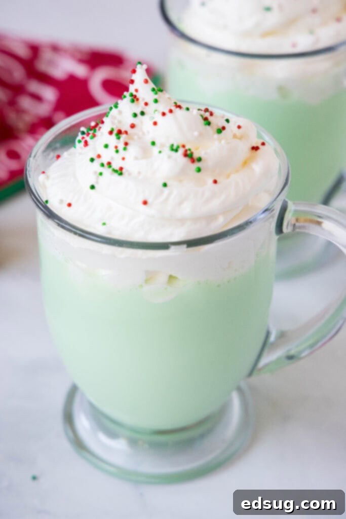 Grinchy Hot Chocolate 2 A mug of creamy green Grinch Hot Chocolate topped generously with whipped cream and festive red and green sprinkles, perfect for holiday cheer.