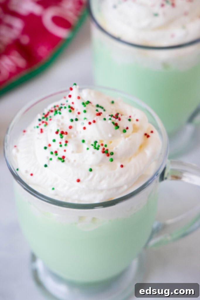 Grinchy Hot Chocolate 6 Close-up of a festive peppermint white hot chocolate in a mug, topped with whipped cream and cheerful red and green Christmas sprinkles.