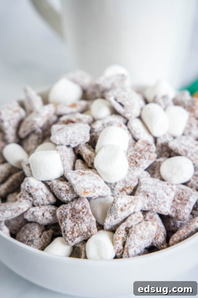 Cozy Cocoa Muddy Buddies 5 A white bowl brimming with hot cocoa muddy buddies and marshmallows, ready for serving