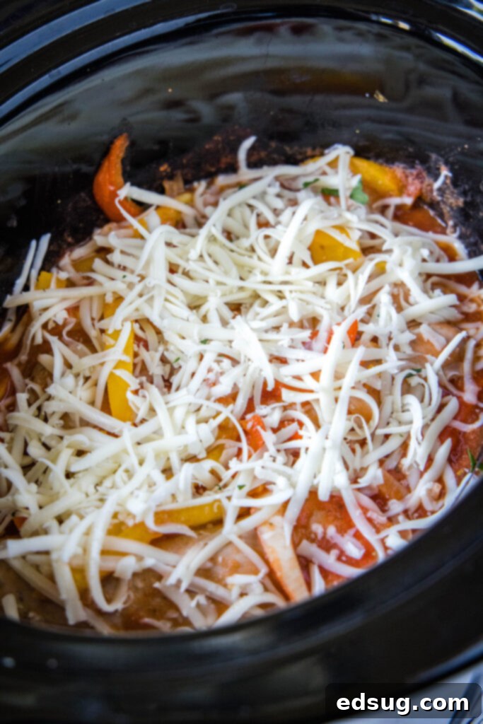 A generous layer of shredded mozzarella cheese melting over the top of hot sausage and peppers in a slow cooker, indicating the final step.