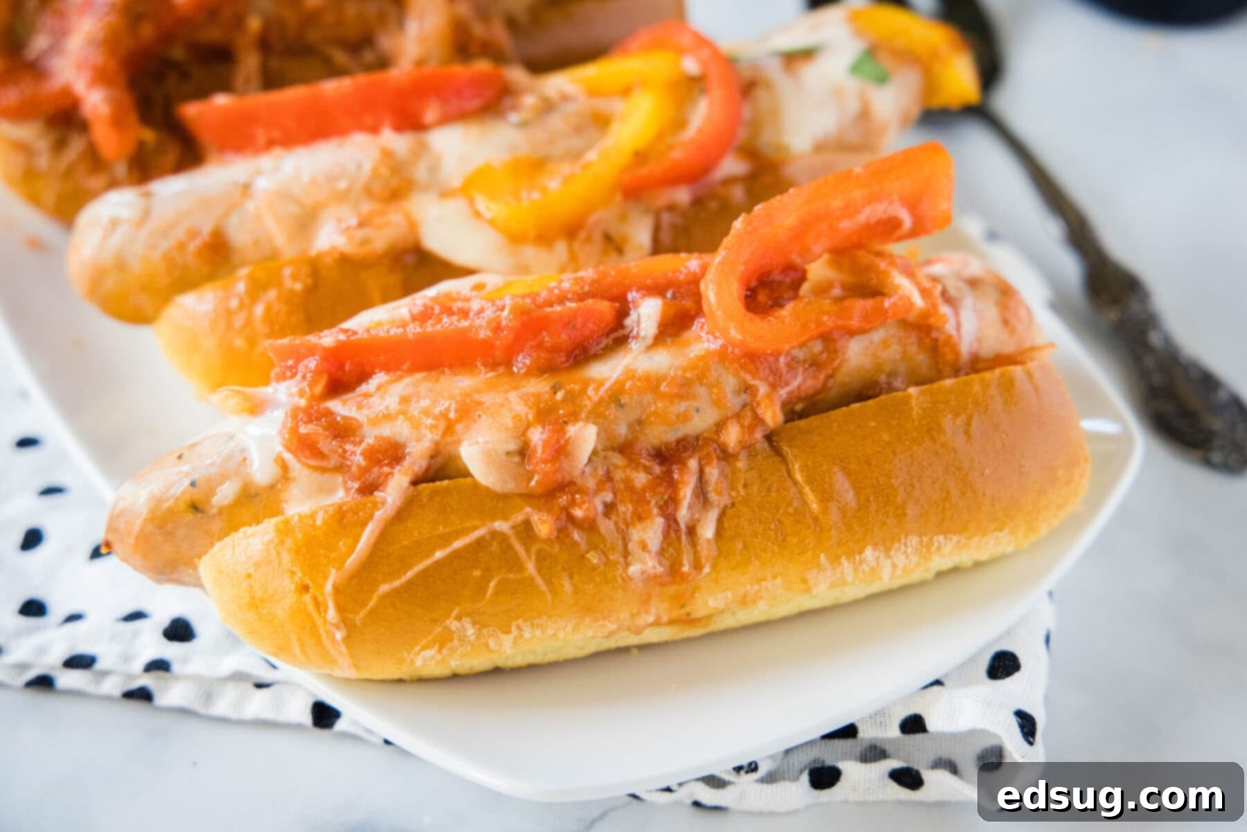 Two perfectly assembled sausage and peppers sandwiches, presented on a plate atop a striped kitchen towel, ready to be eaten.