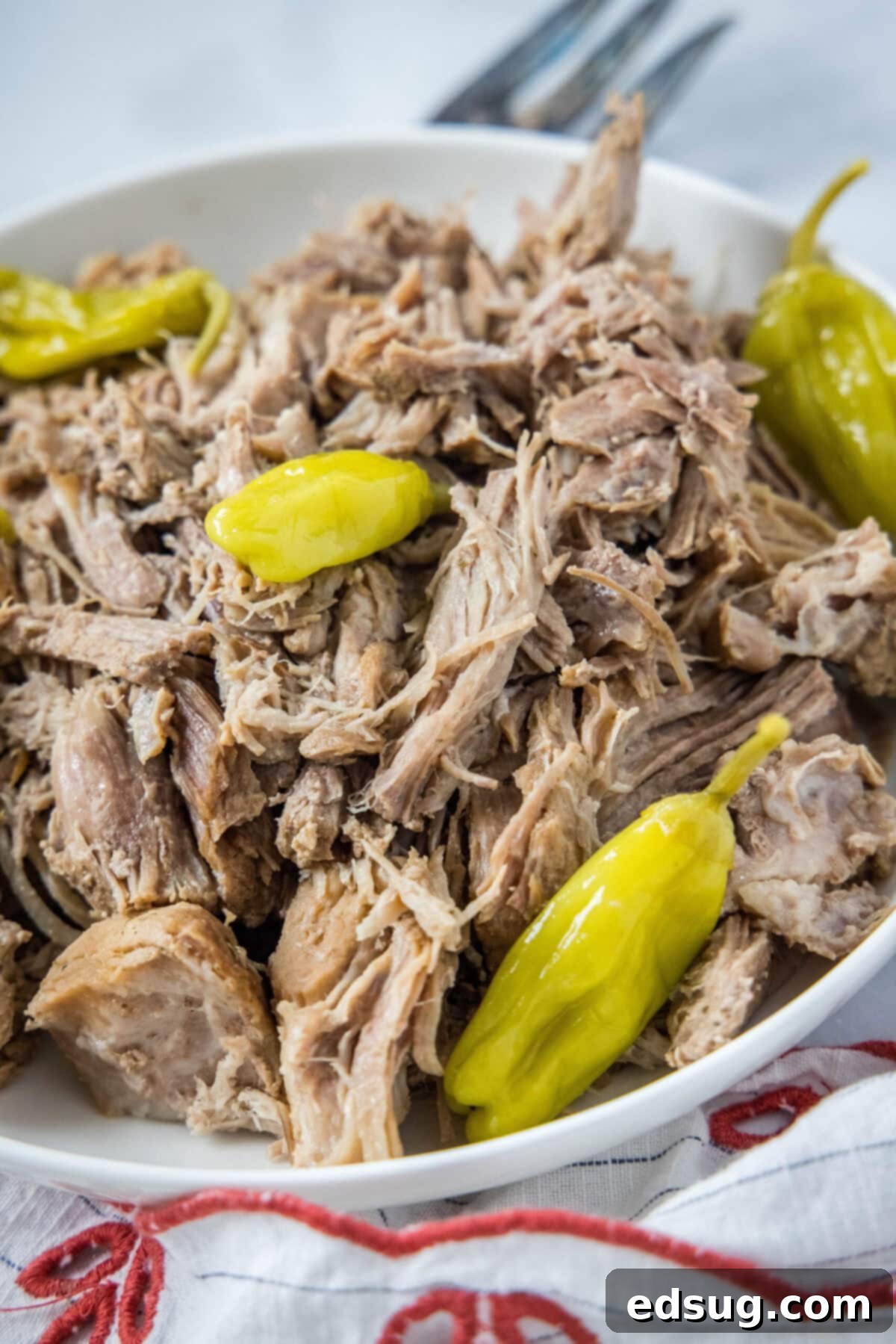 A bowl of shredded pork with pepperoncini peppers, next to a fork