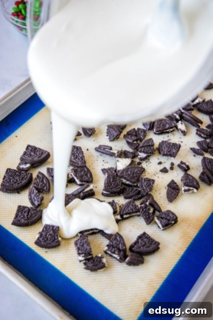 Holiday Candy Bark 4 Melting white chocolate being poured smoothly over a bed of crushed Oreo cookies on a parchment-lined baking sheet.