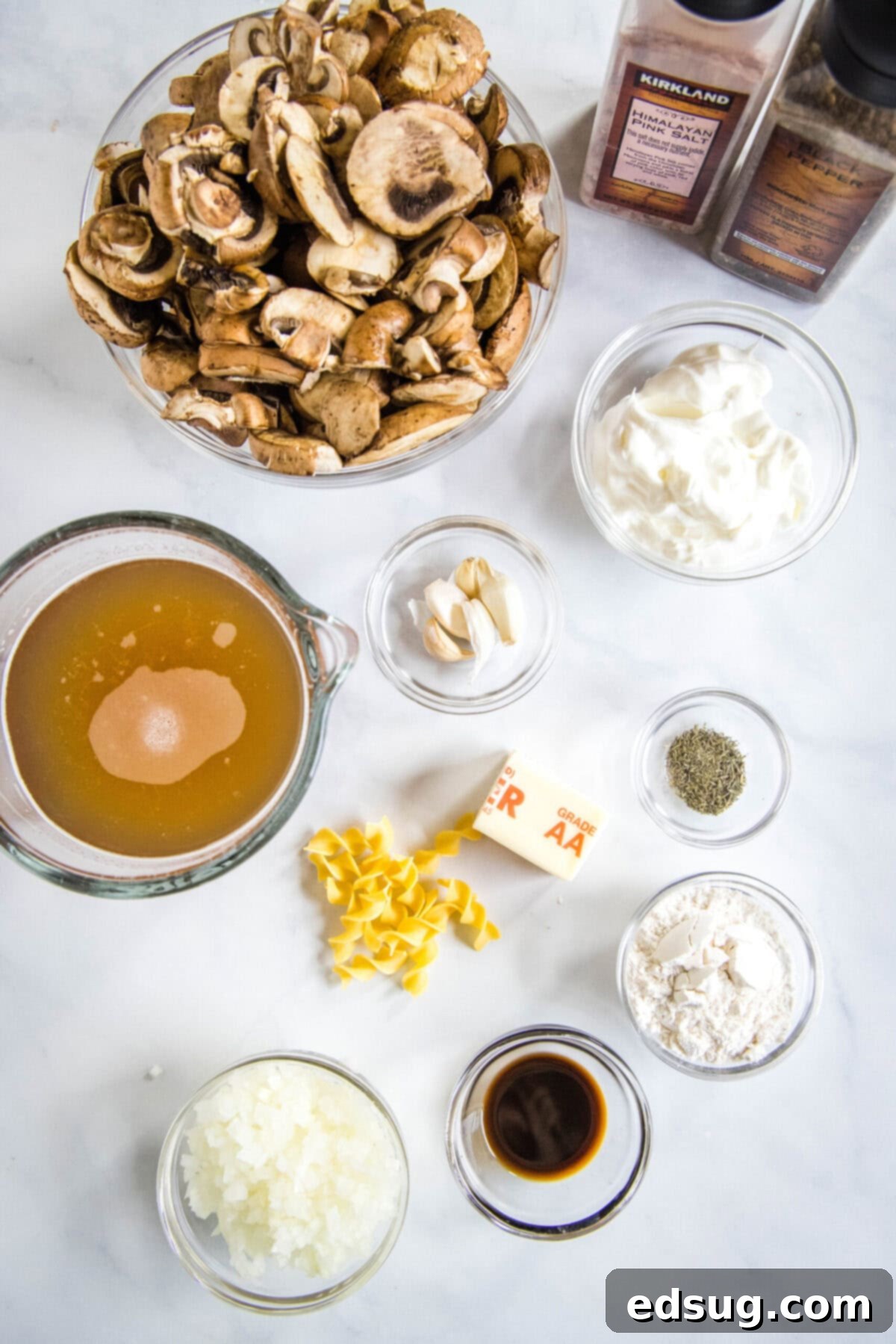 Easy and comforting mushroom stroganoff is loaded with a creamy garlic sauce full of hearty mushrooms, and served over buttery noodles. Overhead view of all the essential ingredients laid out for preparing mushroom stroganoff: a bowl of perfectly sliced mushrooms, a clear Pyrex containing savory broth, a dollop of fresh sour cream, minced garlic, all-purpose flour, finely chopped onions, a bottle of Worcestershire sauce, half a stick of butter, dried thyme, uncooked egg noodles, and salt and pepper grinders for seasoning.