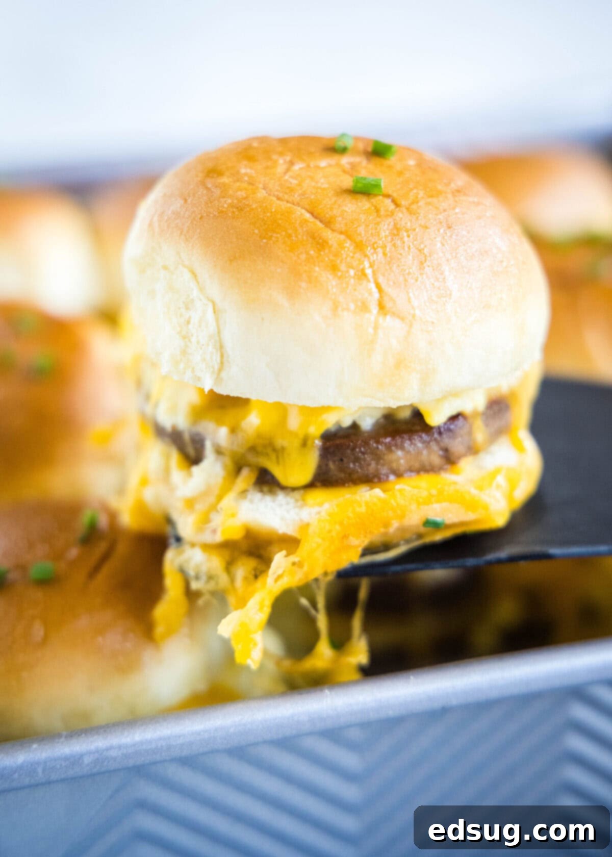 A spatula removing a golden-brown breakfast slider from a baking tray, showing melted cheese and visible layers of egg and sausage.