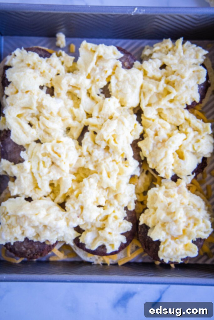 Fluffy scrambled eggs spread over sausage patties and cheese on slider buns in a baking dish.