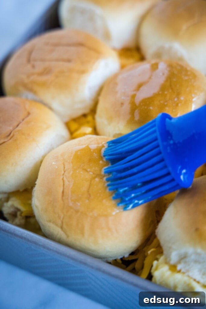 Melted butter and maple syrup being brushed onto the tops of assembled breakfast slider buns in a baking tray.