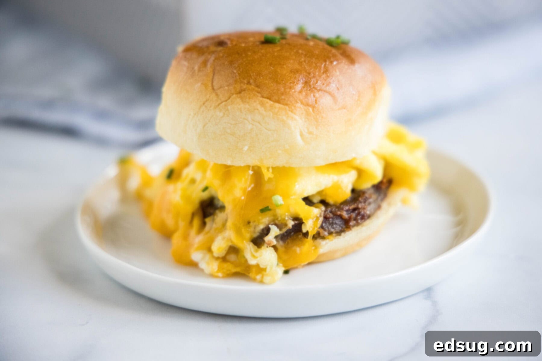 A single breakfast slider on a plate, cut in half to show the layers of sausage, scrambled egg, and deliciously melted cheddar cheese, with a golden-brown bun top.