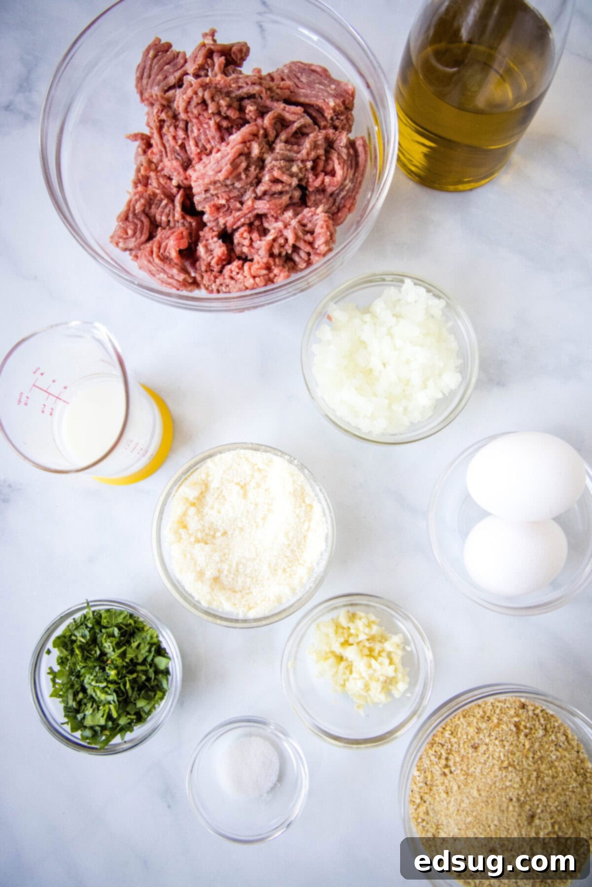 Overhead view of the ingredients needed for air fryer meatballs: a bowl of ground beef, a bowl of onions, a bowl of garlic, a bowl of parsley, a bowl of breadcrumbs, a glass of milk, a bottle of olive oil, a bowl of parmesan cheese, a bowl of salt, and some eggs.
