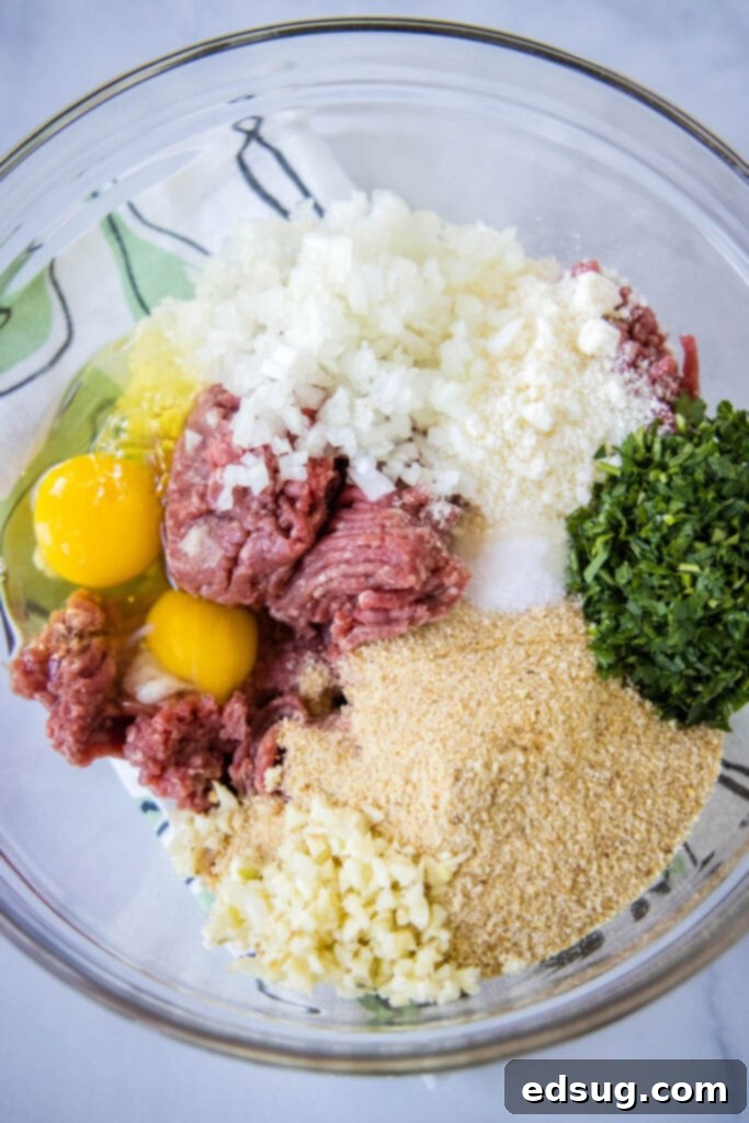 Overhead view of a mixing bowl with ground beef, onions, garlic, salt, breadcrumbs, parsley, eggs, milk, and olive oil.