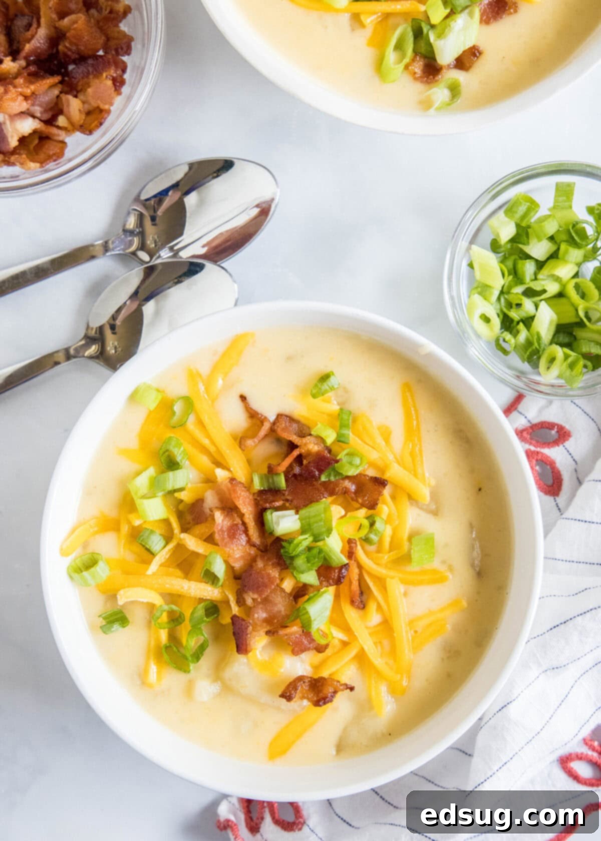With a creamy broth, hearty chunks of potatoes, gooey cheese, and crispy bacon, this Instant Pot potato soup is the ultimate comfort meal. Overhead view of a bowl of potato soup topped with cheese, bacon, and green onions, next to two spoons, a bowl of green onions, a bowl of bacon, and another bowl of soup.