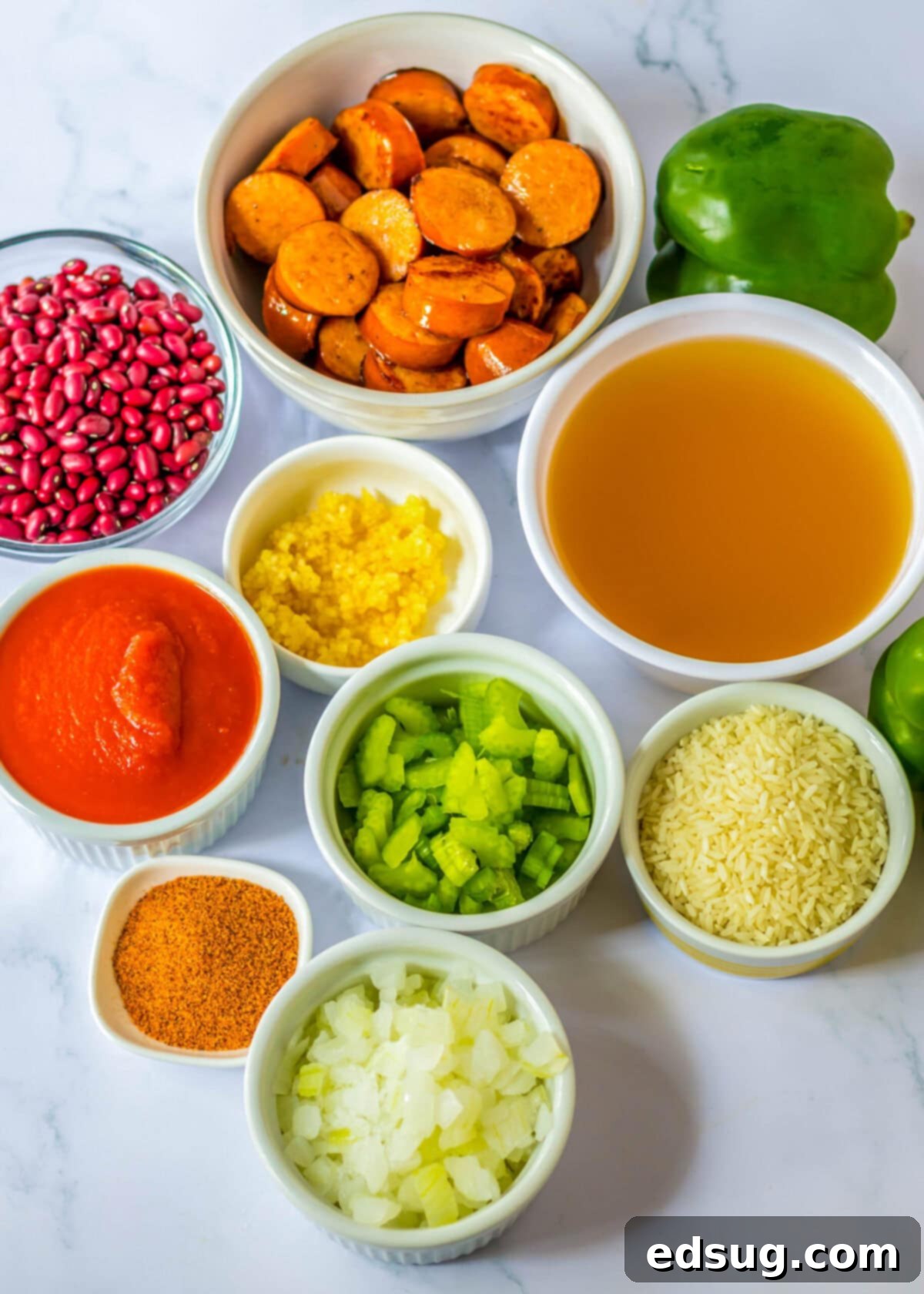 An overhead shot showcasing all the fresh and prepared ingredients for slow cooker red beans and rice: sliced sausage, dried red beans, tomato sauce, chicken broth, diced celery, diced onion, minced garlic, cooked rice, Cajun seasoning, and a whole green bell pepper.