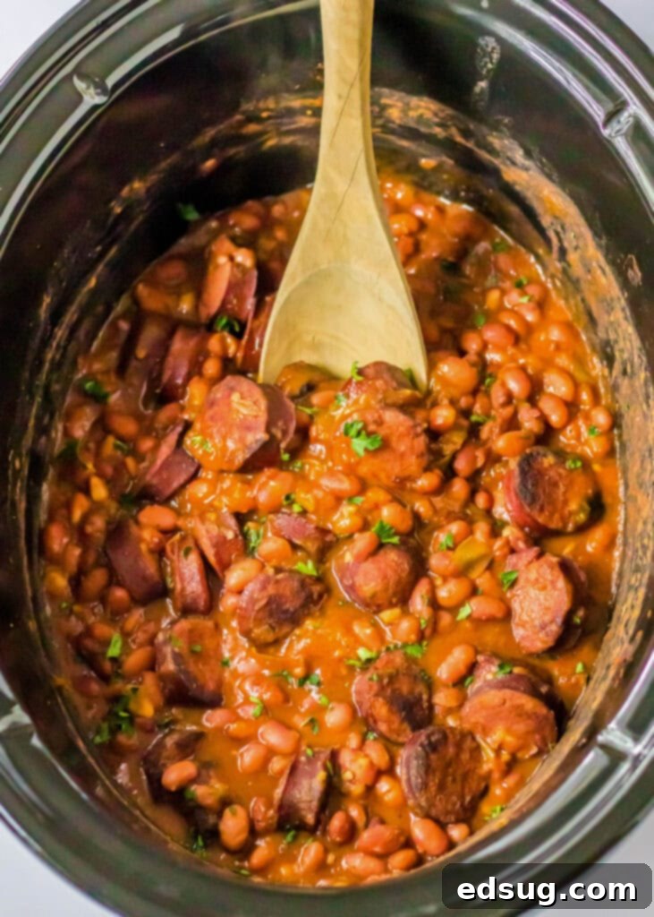 An overhead view of rich, cooked red beans and rice in a slow cooker, with a wooden spoon resting inside, showing the tender beans and sausage.