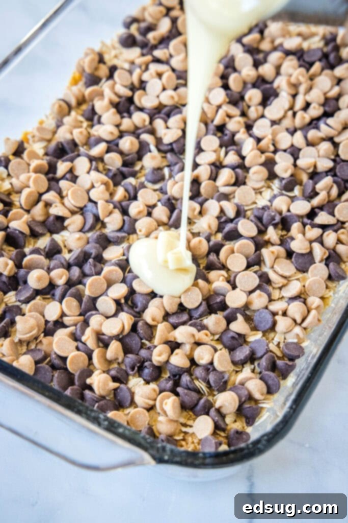 Sweetened condensed milk being poured evenly over a layer of peanut butter and chocolate chips, preparing Hello Dolly bars for baking.