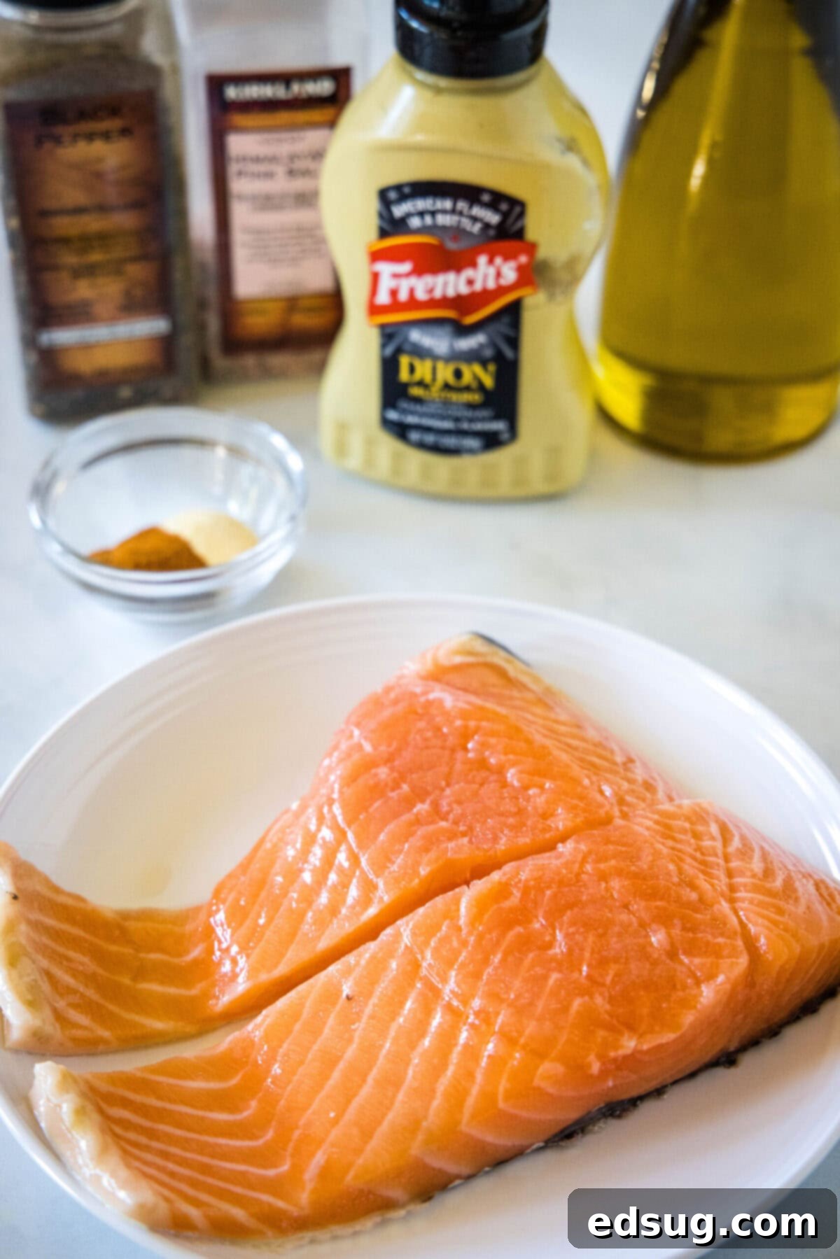 All the ingredients needed to make air fryer salmon: a plate with two salmon fillets, a bowl with garlic powder and paprika, a bottle of Dijon mustard, a bottle of olive oil, and salt and pepper grinders.