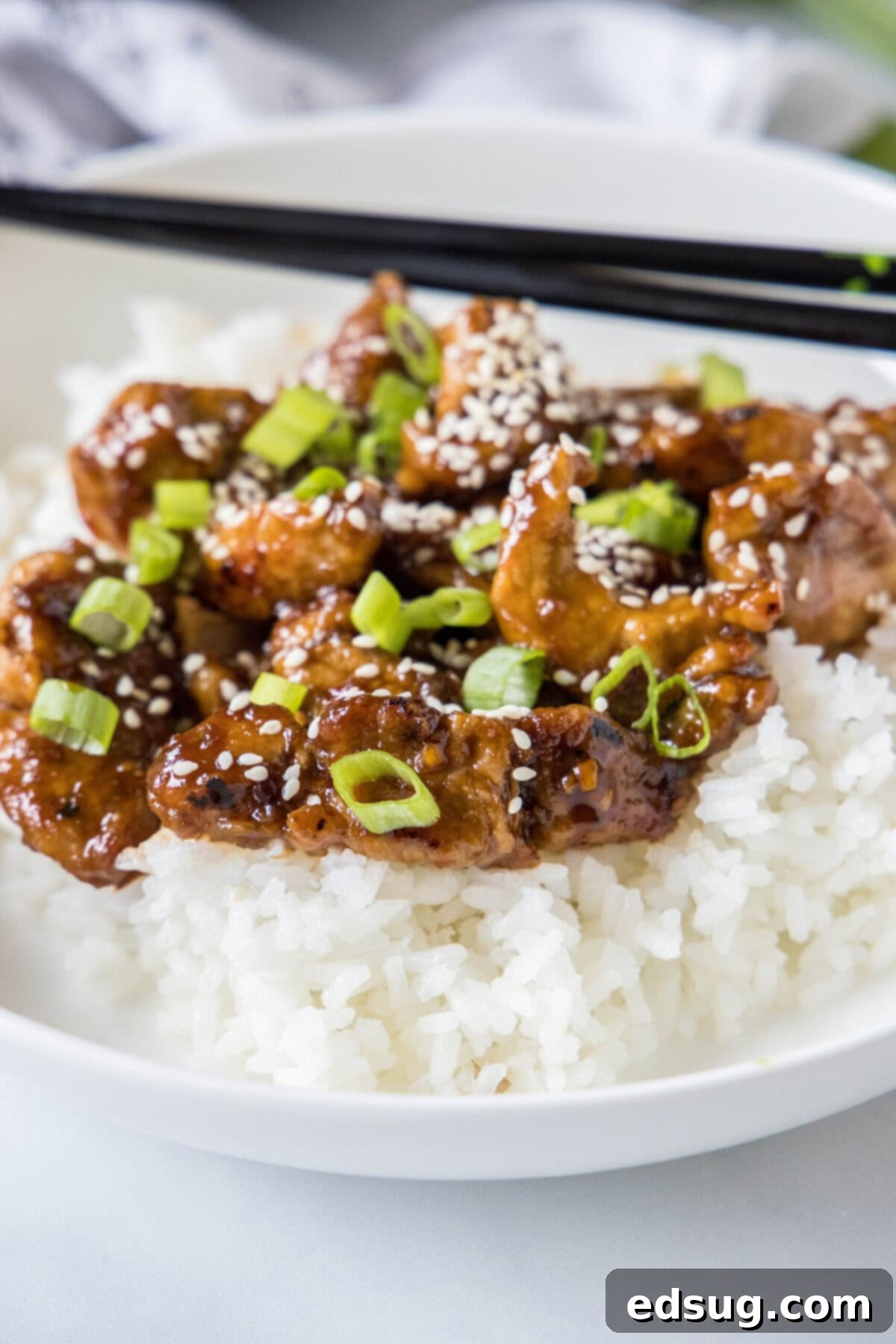 This Mongolian pork is tender, juicy, sweet, sour, a little bit spicy, and so much better than takeout, plus it only takes 20 minutes. Close up of a bowl of rice and Mongolian pork, topped with green onions and sesame seeds, with a pair of chopsticks in the background.