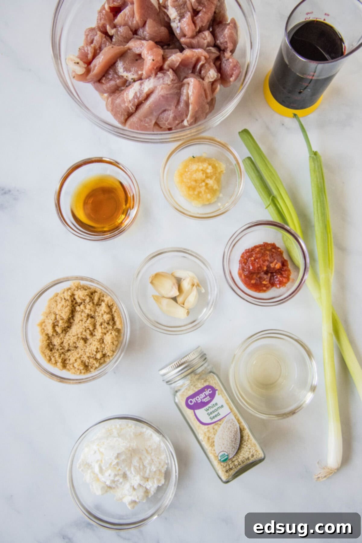This Mongolian pork is tender, juicy, sweet, sour, a little bit spicy, and so much better than takeout, plus it only takes 20 minutes. Overhead view of the ingredients needed for Mongolian pork: a bowl of pork strips, a glass of soy sauce, a glass of vinegar, a bowl of ginger, a bowl of garlic, a bowl of cornstarch, a bowl of brown sugar, a bowl of sesame oil, a jar of sesame seeds, a bowl of chili paste, and two green onions.