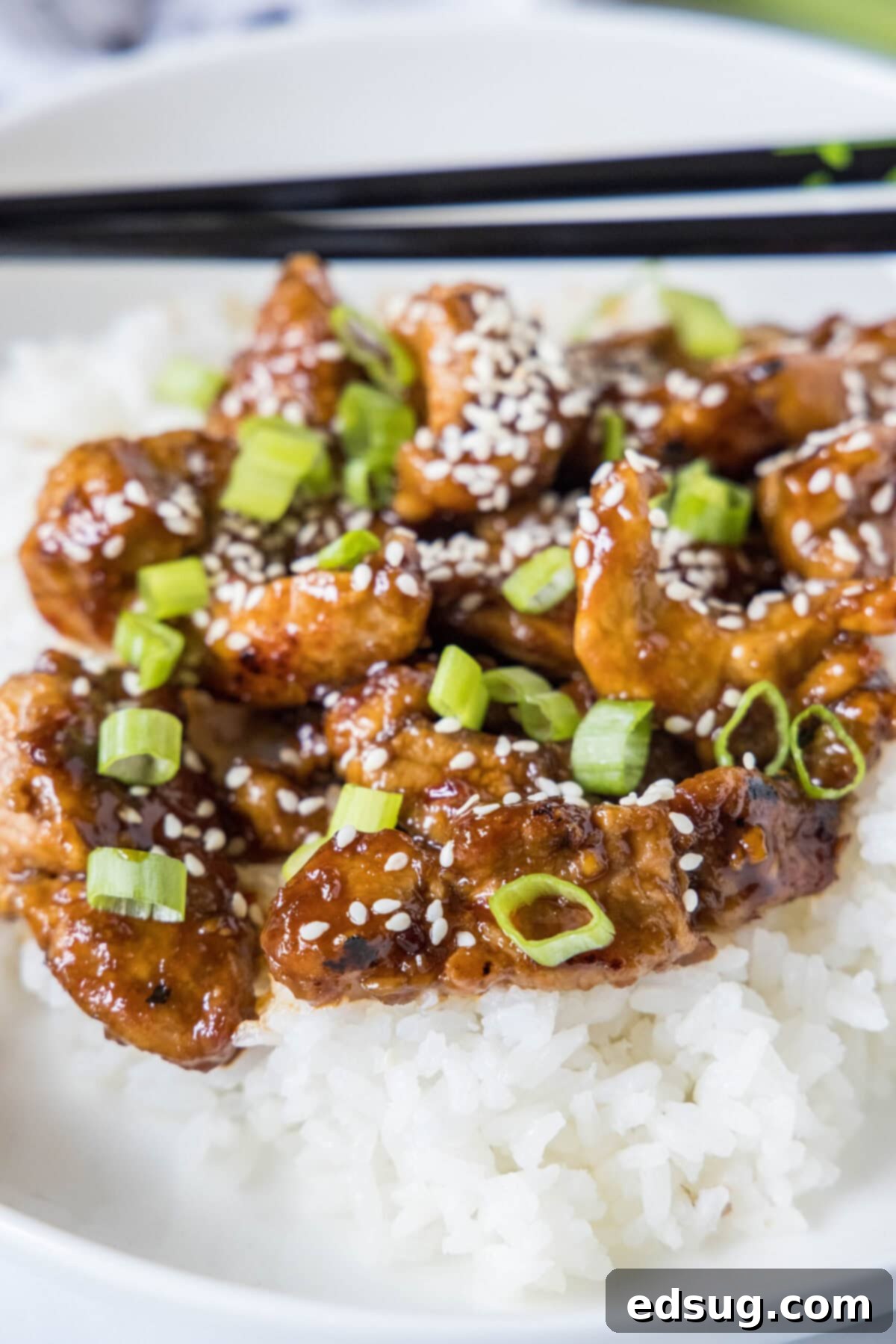 This Mongolian pork is tender, juicy, sweet, sour, a little bit spicy, and so much better than takeout, plus it only takes 20 minutes. Close up of Mongolian pork on top of rice on a plate, topped with green onions and sesame seeds, with a pair of chopsticks.