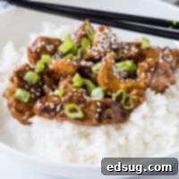 Sweet and Savory Mongolian Pork 2 A plate with Mongolian pork on top of rice, topped with sesame seeds and green onions, with a pair of chopsticks.