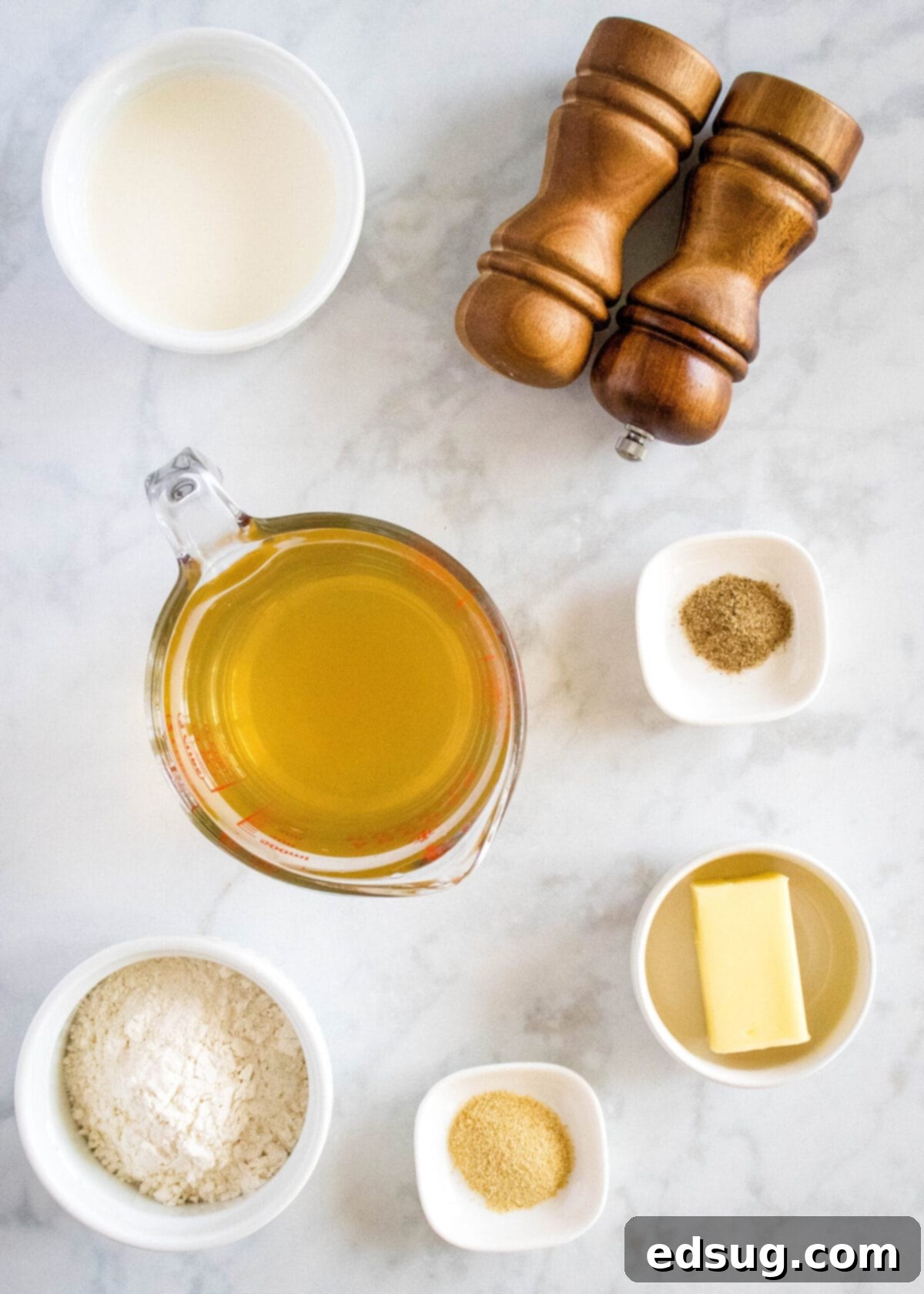 Overhead view of the essential ingredients for homemade cream of chicken soup, neatly arranged: a clear bowl of golden chicken broth, a small pitcher of heavy cream, a bowl of all-purpose flour, a cube of butter, and small bowls containing onion powder, celery salt, and a salt and pepper grinder. These simple ingredients combine to create a rich and easy soup.