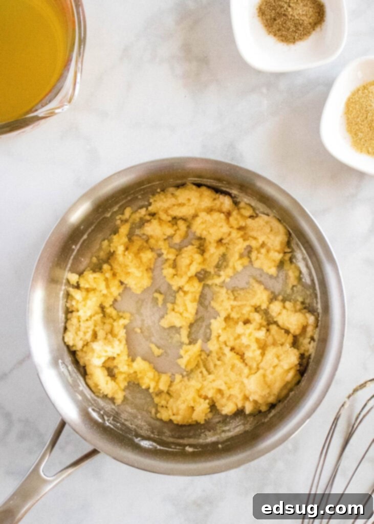 Overhead view of a saucepan on the stove containing a golden roux (a cooked mixture of butter and flour) next to a clear Pyrex measuring cup filled with golden chicken broth, ready to be slowly added and whisked for the soup base.