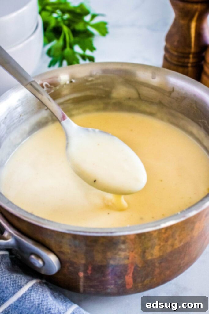 A saucepan filled with a freshly prepared, creamy white cream of chicken soup, with a serving spoon hovering above it, scooping up a portion of the perfectly smooth soup, and fresh parsley in the soft-focus background, ready for plating.