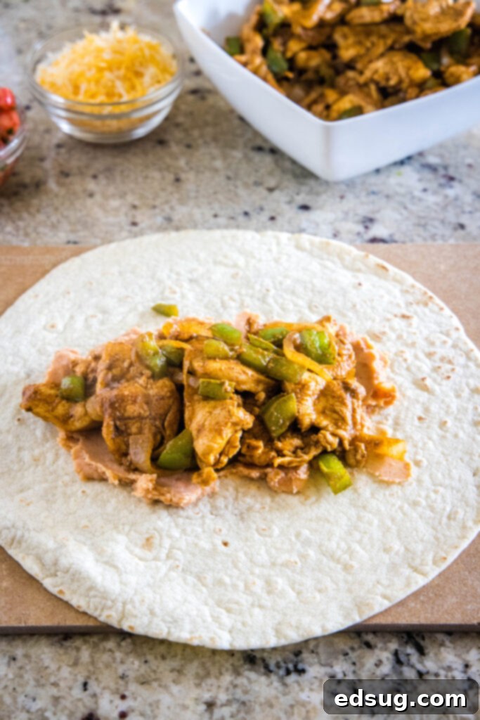 Easy chicken fajita wraps have juicy meat, tender veggies, spicy and smoky seasonings, rich beans, shredded cheese, and fresh salsa. A tortilla topped with beans and chicken fajita mix, next to a bowl of chicken fajitas and a bowl of cheese.